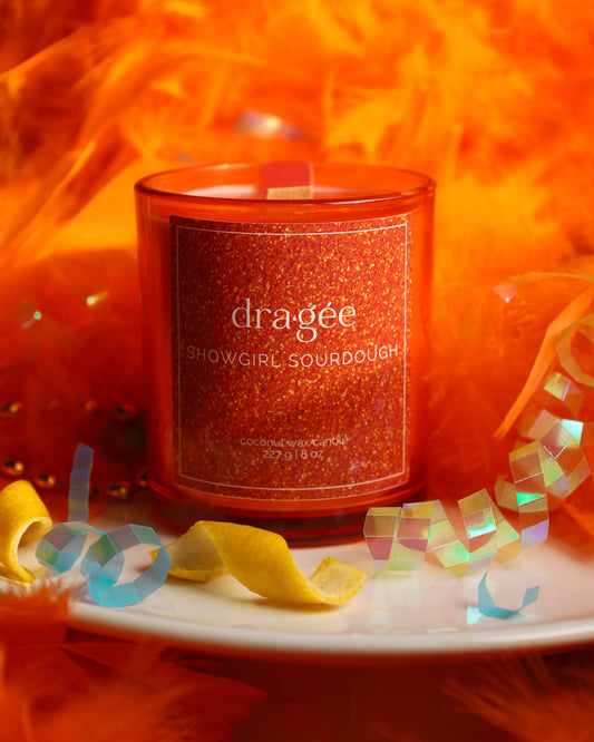 Showgirl Sourdough 8oz Taylor Swift inspired candle blueberry lemon sourdough housed in a vibrant orange glass jar with sparkling label surrounded by an orange feather boa, lemon, and party accessories.