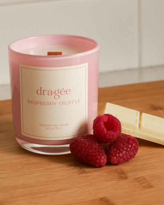 Pink candle with white label says raspberry truffle candle from Dragee Candle Company. Candle is on a cutting board in a kitchen, with raspberries and white chocolate around the candle.
