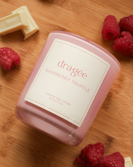 Pink candle with white label says raspberry truffle candle from Dragee Candle Company. Candle is on a cutting board in a kitchen, with raspberries and white chocolate around the candle.