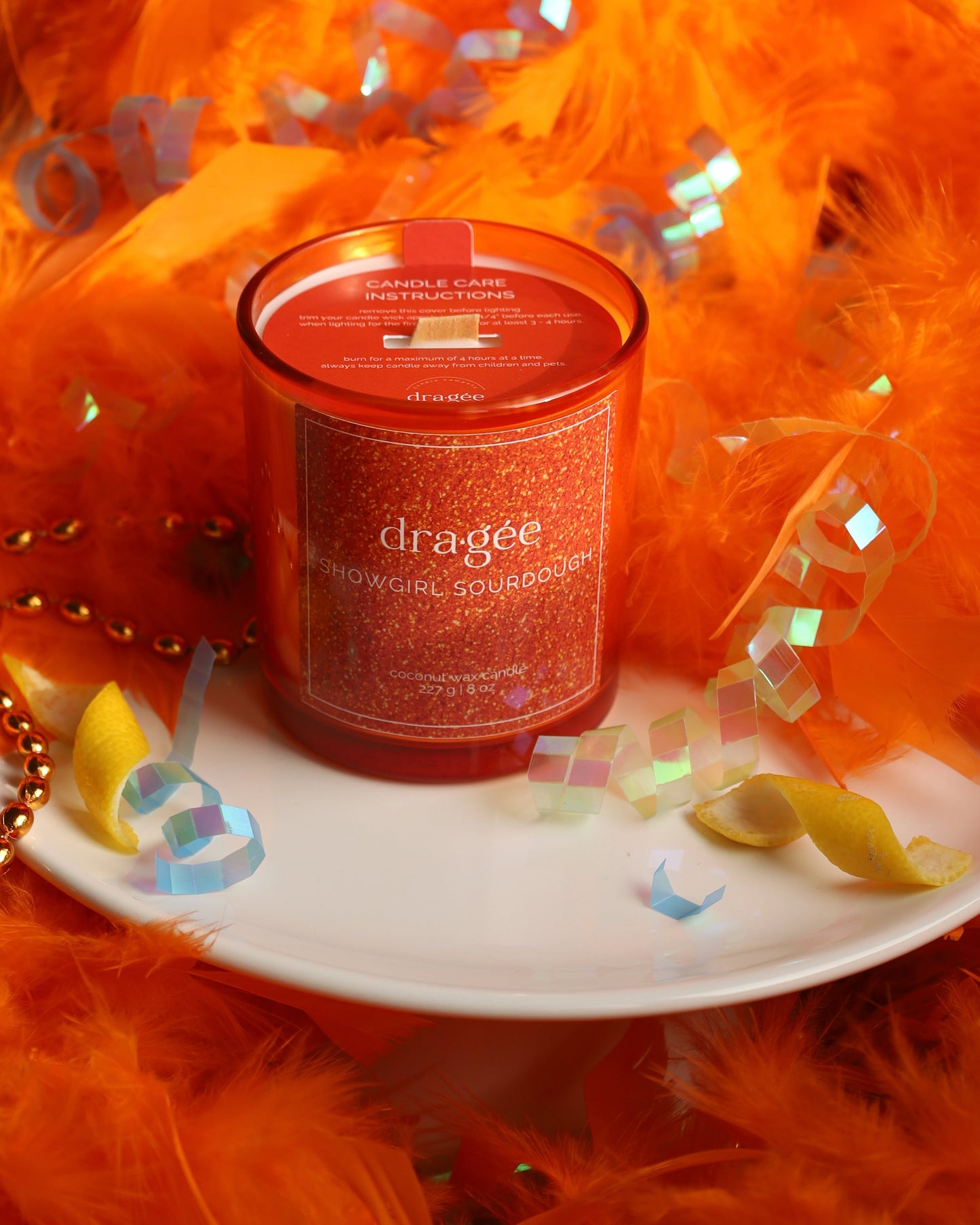 Showgirl Sourdough 8oz Taylor Swift inspired candle blueberry lemon sourdough housed in a vibrant orange glass jar with sparkling label surrounded by an orange feather boa, lemon, and party accessories. 